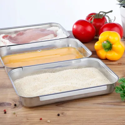 Three stainless steel trays are shown, one with a piece of raw meat dusted in flour, another with beaten eggs, and the third with breadcrumbs. In the background, there are tomatoes and a yellow bell pepper, suggesting ingredients for cooking.