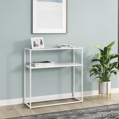 A white console table with two shelves is shown against a light blue wall. There are two framed pictures and a book on the top shelf, and a stack of books on the bottom shelf. A potted plant is to the right of the table.