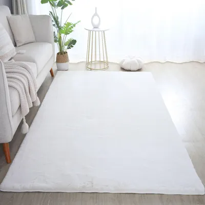 A white fluffy rug is laid out on a wooden floor in front of a sofa. A plant and a small side table are also visible.