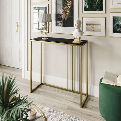 A console table with a black glass top and gold metal frame, decorated with a bust and a lamp, placed in a room with artwork on the wall and a green ottoman.