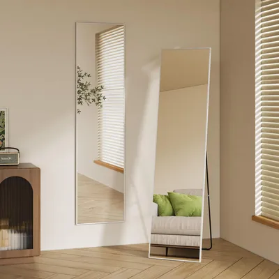 Two full-length mirrors are shown in a room with wooden floors and blinds on the windows. One mirror is leaning against the wall, and the other is standing on a black metal frame. A sofa with green cushions is reflected in the standing mirror.