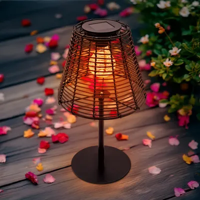 A solar-powered table lamp with a woven shade sits on a wooden deck, surrounded by scattered flower petals and greenery.