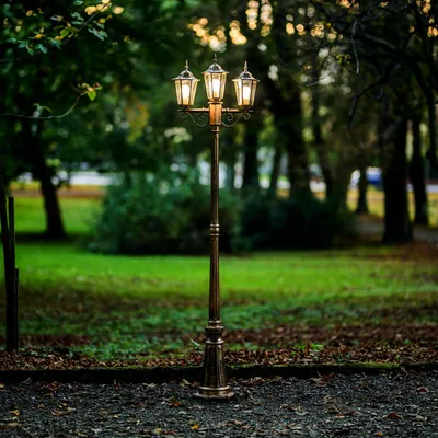 A vintage-style, three-arm lamppost stands in a park-like setting with green grass and trees in the background.