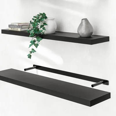 Two black floating shelves are shown mounted on a white wall. The top shelf has books, a white vase with a plant, and a grey vase. The bottom shelf is shown with its mounting brackets.