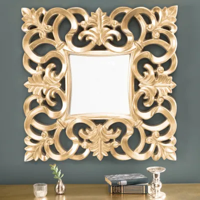 A gold baroque-style mirror is displayed on a dark gray wall above a wooden surface with decorative items.