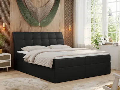 A bedroom scene featuring a black boxspring bed with a tufted headboard, set against a wood-paneled wall with macrame wall art. A window with sheer curtains and plants is visible on the right.