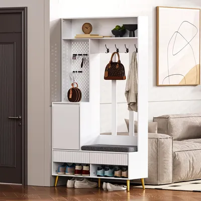 A white entryway storage unit with a bench, shoe rack, cabinet, and hooks, placed in a home setting next to a door and a sofa.