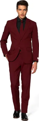 A man wearing a burgundy suit and black shirt and tie, standing against a white background.