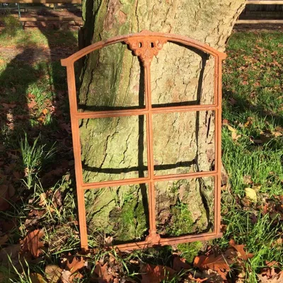 An antique cast iron window frame leaning against a tree trunk in an outdoor setting.