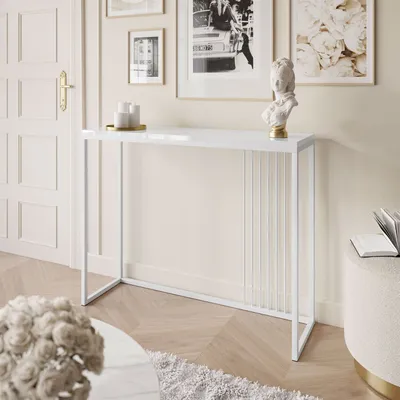 A white console table with a glossy finish is placed in a well-lit room, next to a white door and framed artwork on the wall. Decorative items like candles and a bust are placed on the table.