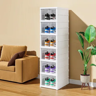 A stack of white shoe storage boxes filled with various pairs of sneakers, placed next to a sofa and a potted plant.