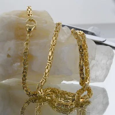 A gold chain necklace is displayed on a light-colored, textured surface, with a reflection visible below.