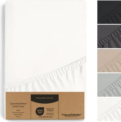 A white fitted sheet is shown folded and partially unfolded, with a cardboard tag attached to the bottom corner. Several other colors of the fitted sheet are displayed in small stacked images on the right side.