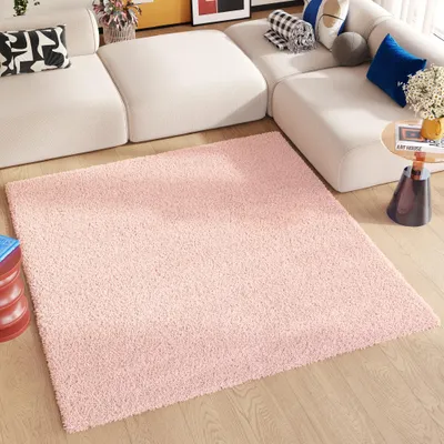 A pink rug is placed on a wooden floor in front of a white sectional sofa. Decorative pillows and a small side table with a vase of flowers are visible on the sofa.