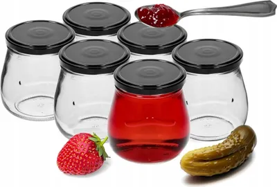 A set of six empty glass jars with black lids, one of which is filled with a red liquid. A strawberry and a pickle are placed in front of the jars.