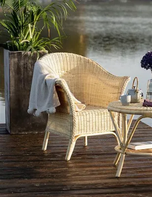 A wicker chair and small table are set up on a wooden deck next to a body of water, with a plant in a planter.