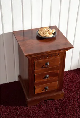A wooden nightstand with three drawers is placed against a white paneled wall and on a dark red carpet.