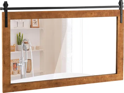 A rectangular mirror with a wooden frame, designed to look like a barn door, is mounted on a wall above a bathtub. The mirror reflects a bathroom shelf with a plant and shower fixtures.