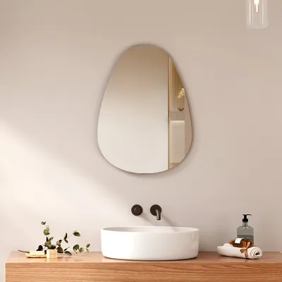 A modern bathroom vanity with a sink, faucet, and an asymmetrical mirror above it. A small plant and a soap dispenser are also visible.