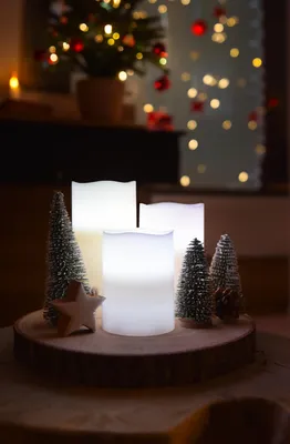 A festive Christmas arrangement with LED candles and miniature trees on a wooden slice, with a blurred Christmas tree and lights in the background.