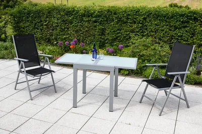 A patio set with two chairs and a table on a tiled patio, with a hedge and flowers in the background.