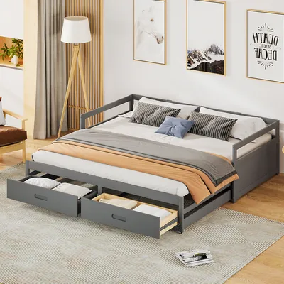 A grey daybed with two drawers pulled out, showing bedding inside. The daybed is in a room with artwork on the wall and a floor lamp.