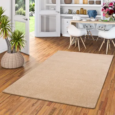 A beige rug is placed on a wooden floor in a dining room setting, with a dining table and chairs visible in the background.