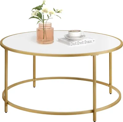 A round coffee table with a white top and a gold metal frame, decorated with a vase of flowers and a book with a cup of coffee on top.