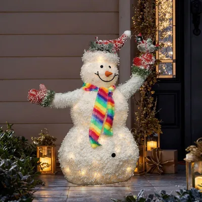 A lighted snowman decoration with a rainbow scarf and a cardinal on its arm, placed on a porch during nighttime.
