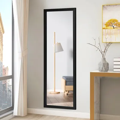 A full-length mirror with a black frame is shown leaning against a light gray wall, next to a window with white curtains. A floor lamp and a bench are reflected in the mirror. A small table with a vase and decorative branches is to the right of the mirror.