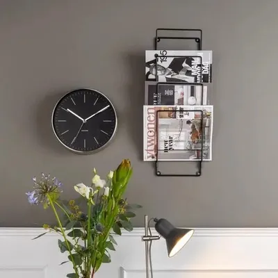 A wall with a clock and a magazine rack, with flowers and a lamp in the foreground.