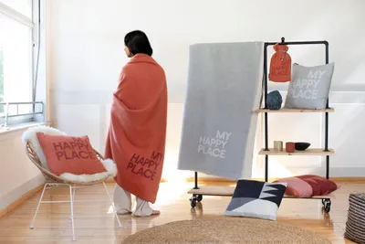 A person wrapped in a coral blanket stands in a room with various home decor items, including pillows and a shelf unit.