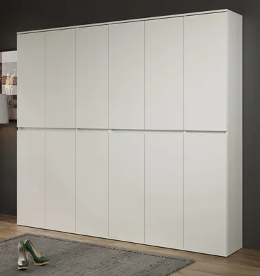 A large white wardrobe with multiple doors, shown in a room with a dark gray wall and a wooden floor. A grey rug is in front of the wardrobe, with a pair of green high-heeled shoes on it.