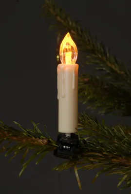 A close-up of a white LED candle light with a yellow-orange flame, clipped onto a green pine branch.