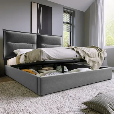 A modern bedroom with a grey upholstered bed frame featuring a lift-up storage base. The bed is made up with white and beige bedding.