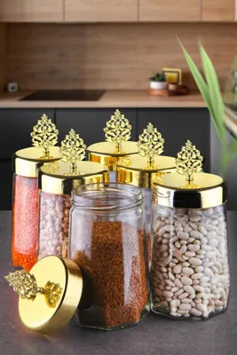 A set of six glass storage jars with decorative gold lids, filled with various dry goods, arranged on a kitchen counter.