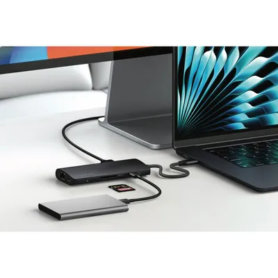 A Satechi USB-C multi-port adapter is connected to a laptop, with an external hard drive and an SD card also connected to the adapter. A large monitor is visible in the background.
