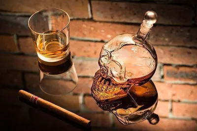 A glass skull decanter filled with amber liquid, a matching glass with amber liquid, and a cigar are arranged on a reflective surface in front of a brick wall.