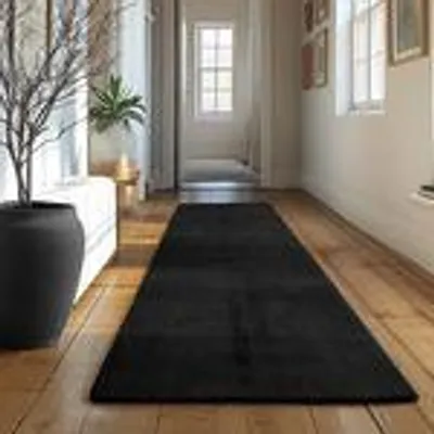 A black rug is laid out on a wooden floor in a hallway, with a large black vase and a plant on the left.