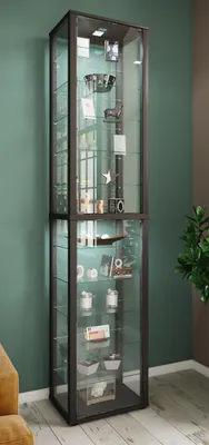 A tall, narrow glass display cabinet with multiple shelves, filled with various decorative items, standing against a green wall.