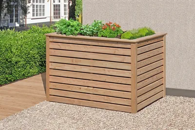 A wooden planter box filled with plants sits on a gravel and wooden deck area next to a building.