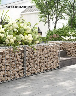 A SONGMICS gabion basket filled with stones and topped with white flowers, set in an outdoor garden setting with a house in the background.