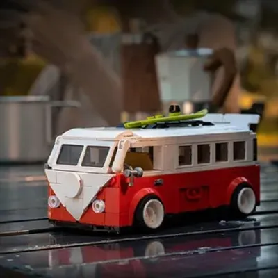 A Lego-style camping bus on a wooden surface, with blurred background elements suggesting a campsite or kitchen setting.