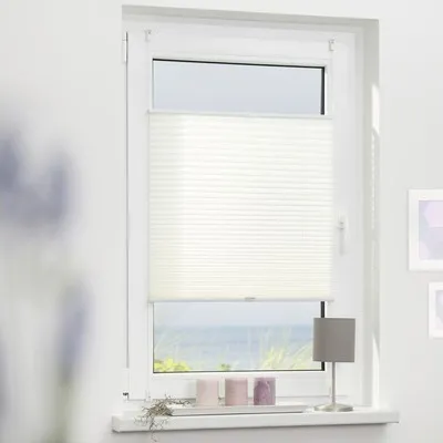 A white pleated blind is installed on a window overlooking a beach scene. There are three candles and a small lamp on the windowsill.