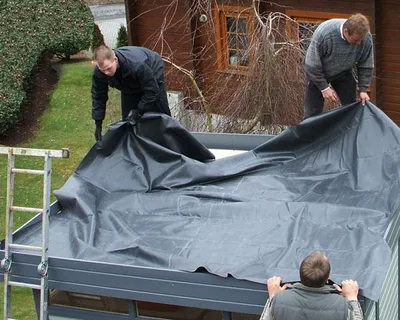 Two men are installing a large black tarp on a roof.