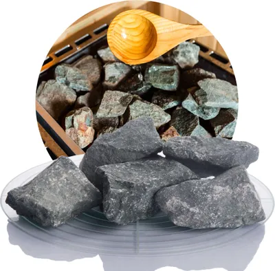 A pile of dark gray sauna stones on a clear plate, with more stones and a wooden scoop visible in the background within a circular frame.