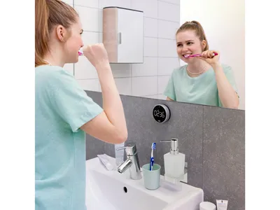 A young woman brushing her teeth in front of a bathroom mirror, with a digital timer attached to the mirror.