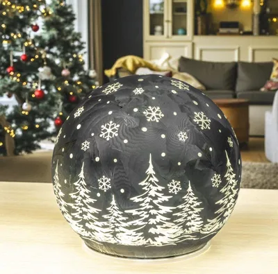 A decorative LED sphere with a Christmas tree and snowflake design sits on a table in front of a decorated Christmas tree and a living room setting.