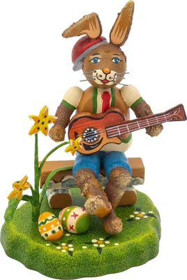 Ein fröhlicher Hase mit roter Mütze und Gitarre sitzt auf einer Bank umgeben von bunten Ostereiern und gelben Blumen auf einer grünen Wiese