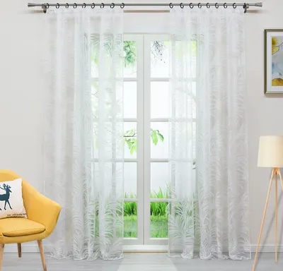A bright room with a window covered by white sheer curtains with a leaf pattern. A yellow armchair with a pillow is in the foreground.